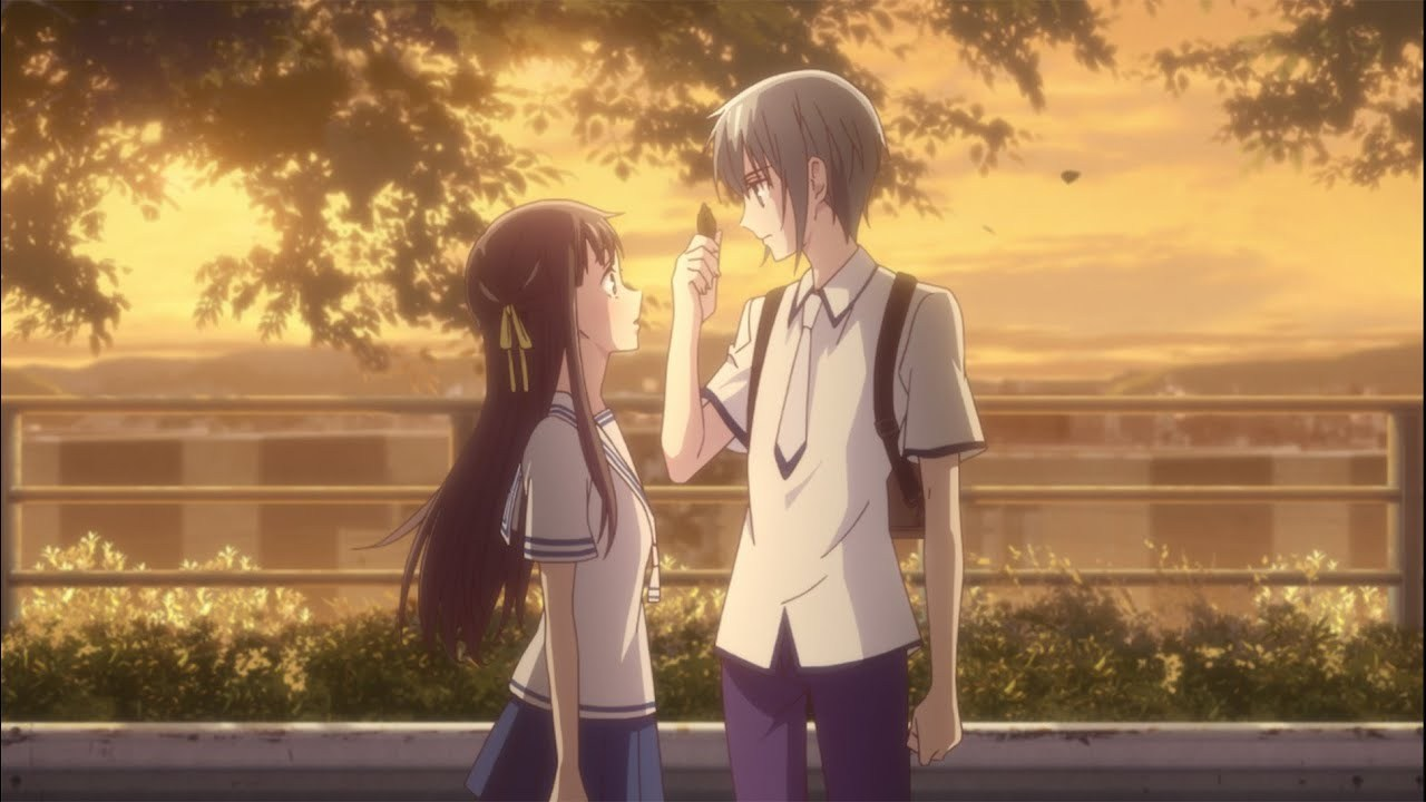 Cena de Fruits Basket ao entardecer: dois estudantes de uniforme, um rapaz de cabelo claro e uma garota de cabelo longo escuro, se encaram em uma rua arborizada com luz dourada; o rapaz segura uma folha entre os dedos, como um gesto delicado de aproximação.