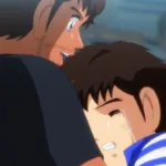 Cena de Captain Tsubasa (Super Campeões) em que Tsubasa, um garoto japonês emocionado e chorando, abraça Roberto, um homem brasileiro, em um estádio de futebol com arquibancadas desfocadas ao fundo. Imagem de Captain Tsubasa (Super Campeões) mostrando um japonês chorando nos braços de um brasileiro: Tsubasa abraça Roberto no estádio. A cena conversa com o clima de Brasil x Japão ao transformar rivalidade e contraste de estilos em algo mais humano — respeito, amizade e a vontade de “mais jogo” quando o apito final não basta.