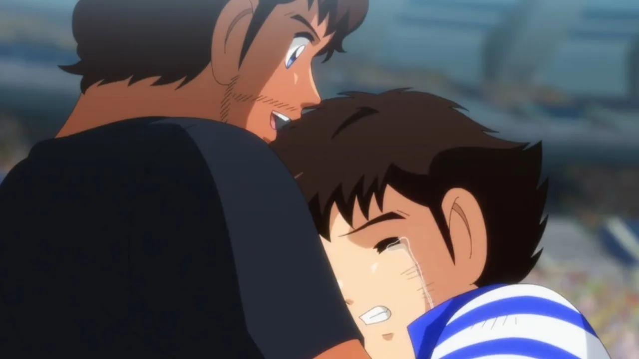 Cena de Captain Tsubasa (Super Campeões) em que Tsubasa, um garoto japonês emocionado e chorando, abraça Roberto, um homem brasileiro, em um estádio de futebol com arquibancadas desfocadas ao fundo. Imagem de Captain Tsubasa (Super Campeões) mostrando um japonês chorando nos braços de um brasileiro: Tsubasa abraça Roberto no estádio. A cena conversa com o clima de Brasil x Japão ao transformar rivalidade e contraste de estilos em algo mais humano — respeito, amizade e a vontade de “mais jogo” quando o apito final não basta.