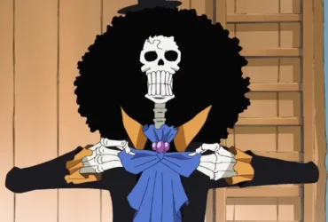 Brook, personagem esqueleto de One Piece, aparece de frente com um grande cabelo afro preto, casaco escuro com detalhes amarelos e laço azul no pescoço. Ele segura as lapelas da roupa em um ambiente de madeira, ao lado de uma cortina azul.