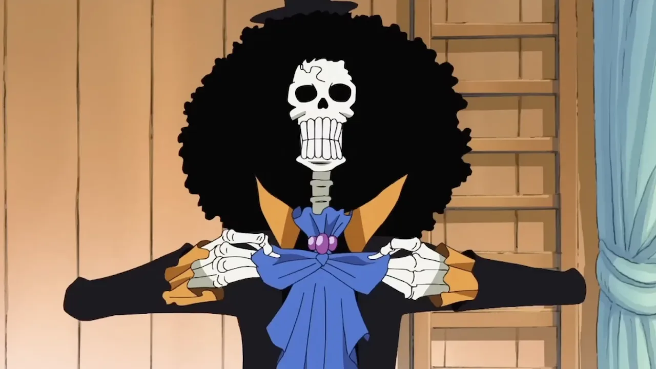Brook, personagem esqueleto de One Piece, aparece de frente com um grande cabelo afro preto, casaco escuro com detalhes amarelos e laço azul no pescoço. Ele segura as lapelas da roupa em um ambiente de madeira, ao lado de uma cortina azul.