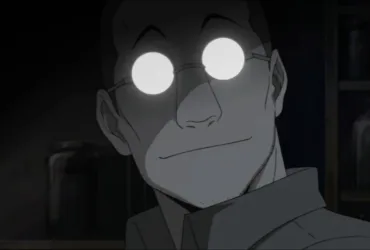 Shou Tucker, personagem de Fullmetal Alchemist, aparece em close em um laboratório escuro. Seus óculos redondos refletem uma luz branca intensa e escondem seus olhos, enquanto ele exibe um sorriso discreto. Ao fundo, prateleiras com frascos reforçam a atmosfera fria, científica e inquietante da cena.