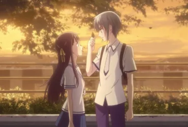 Yuki Sohma e Tohru Honda, personagens de Fruits Basket, estão lado a lado em uniforme escolar ao entardecer. Yuki, de cabelo cinza, segura um pequeno objeto diante de Tohru, que olha para ele. Árvores, folhas ao vento e uma luz dourada de pôr do sol criam uma atmosfera de cuidado, calma e conexão.